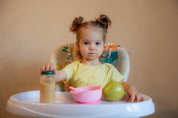 A little girl is sitting on a high chair and eating mashed fruit at home