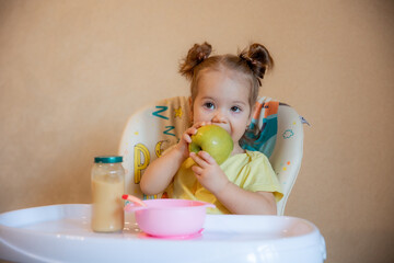 A little girl is sitting on a high chair and eating a green apple