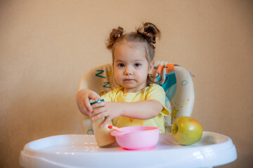 A little girl is sitting on a high chair and eating mashed fruit at home