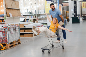Happy couple shopaholics in store, woman sitting on trolley, cheerful husband rolls her in...