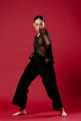 Fashion asian woman in sheer top and black pants posing on red background with hands on hips and legs spread apart