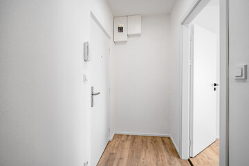 A bright, empty hallway with white walls, a wooden floor, an open door leading to another room, and an intercom system on the wall.