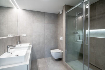 A modern bathroom with gray tiles, a double sink with chrome faucets, a glass-enclosed shower, a wall-mounted toilet, and a large mirror.