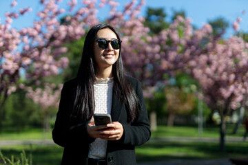 A woman is holding a cell phone in a park with cherry blossoms in the background. She is smiling and she is enjoying her time outdoors