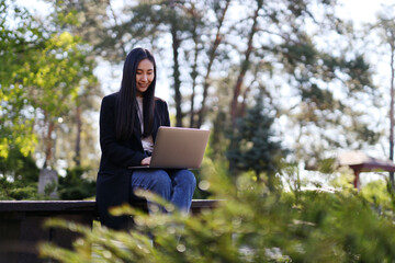 A woman is sitting on a bench with a laptop in front of her. She is wearing a black coat and blue jeans. The scene is set in a park with trees in the background