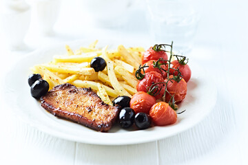 Roast Pork Steak with french fries, tomatoes and olives. White wooden background. Close up.	