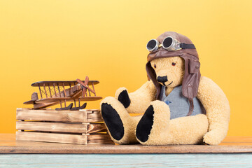 Teddy Bear toy in leather aviator's hat and vintage goggles, wooden plane front yellow wall background. Retro old style filtered photo