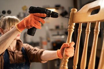 Female craftsman in apron and shirt making holes in chair during repair, standing in her workshop. concept repair and restoration of old furniture