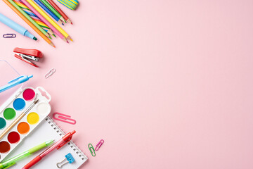 A top view of colorful school stationery like pencils, paints, and notebooks arranged on a pink...