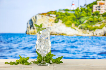 Glass of still mineral water with ice and fresh mint   on wooden board table and rocky seaside and tropical beach background.