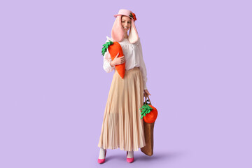 Stylish woman with bunny ears holding carrot toys on lilac background. Easter celebration