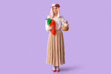 Beautiful young woman in bunny ears and stylish skirt holding toy carrot and basket with Easter bread on lilac background. Easter celebration