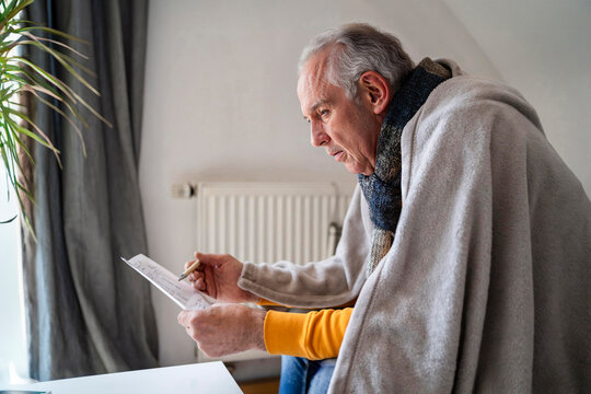 Sick senior man with shawl sitting on sofa and going through medical bill at home