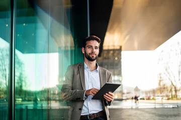 Smiling businessman holding tablet PC and leaning on glass