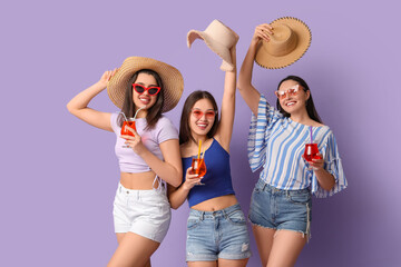 Beautiful young women with glasses of cocktail and wicker hats on lilac background. Summer party