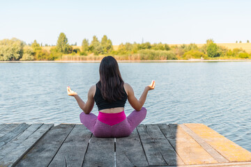 Back view, young sporty woman practicing meditation in lotus pose on wooden pier by a river, morning workout outdoors