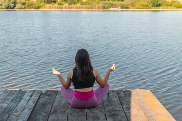 Back view, young sporty woman practicing meditation in lotus pose on wooden pier by a river, morning workout outdoors