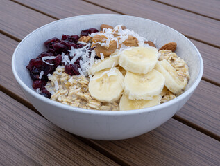 Oatmeal with bananas, cranberries, coconut and peanut butter in a white bowl on wooden table.