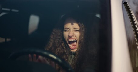 An emotional teenage girl with curly hair and braces screams while gripping the steering wheel of a car, expressing stress or fear.