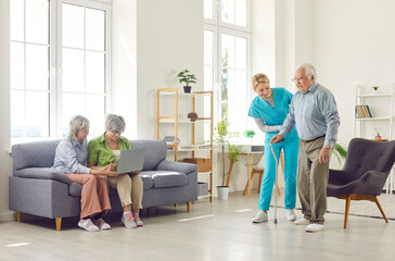 Fototapeta premium Daily lifestyle in retirement home. Retired people spend time in assisted living facility. Young nurse helps old man walk with stick while older ladies sit on sofa with laptop. Senior care concept