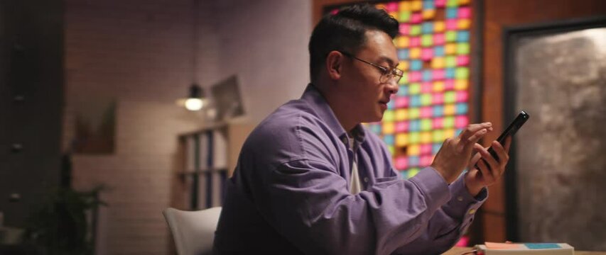 Handsome young asian ethnicity man with glasses use mobile phone scrolling chatting in social media at night evening home. 360 degree. Camera moving around