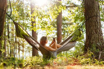 A green hammock hangs among the trees by the lake. A young girl is resting outdoors in a hammock. Picnic. Summer outdoor recreation with an overnight stay. Summer background