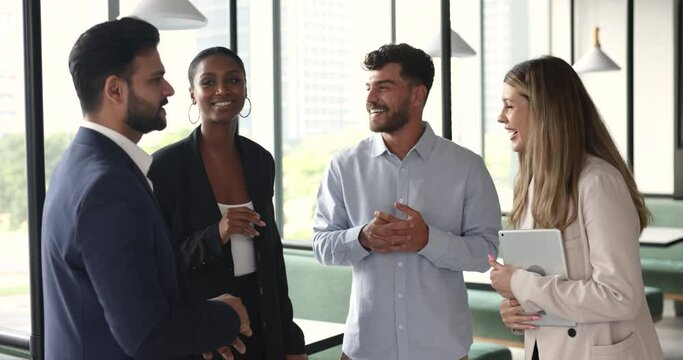 Diverse team of young business colleagues chatting in office hall. Multiethnic office friends discussing corporate life on work break, standing together, talking, smiling, laughing