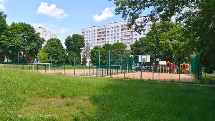 Sandy soccer and basketball courts with metal gates and plastic boards, with poles with lighting lanterns are fenced with a metal mesh fence. Around there are residential houses and trees