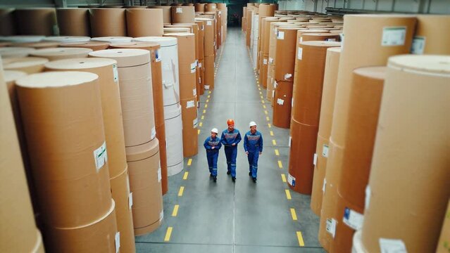 Paper mill workers walk through a huge warehouse with large rolls of paper. Engineers are taking inventory of warehouse. Paper industry, stock. Top view, drone. Factory, plant, engineers, workers.