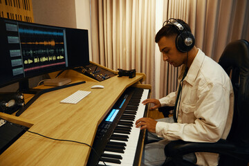 Musician in headphones sitting at his workplace and playing on synthesizer to record song