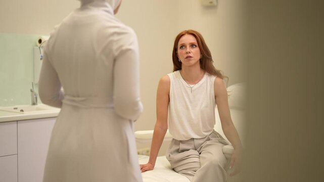 Worried female patient listening to doctor during medical examination in modern clinic. Caucasian young woman sits on examination couch in exam room and listening to advice of female pediatrician.