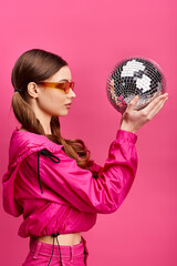 A young stylish woman in her 20s wearing a pink jacket embraces a disco ball in a vibrant studio setting with a pink background.