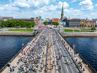 Aerial shot of marathon participants crossing a bridge over a river in Riga, Latvia. The city skyline and St. Peter's Church are visible in the background.
