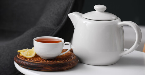 Board with cup of hot brewed tea, slices of lemon and pot on table against black blurred background