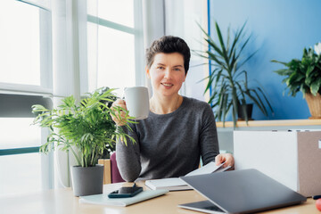 Satisfied smiling newcomer middle aged neutral gender female employee holds coffee cup and sitting at her new office desk work space. First working day concept. Dreaming about new opportunities