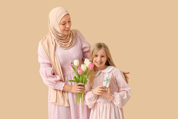 Beautiful young Muslim woman holding bouquet of tulips and her cute little daughter with gift box on beige background