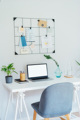 Modern Contemporary workplace design. White desk with laptop mockup empty screen, grid mood board with pinned notes and plans, green plants, office supplies at work space in home office room interior
