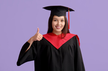 Young female graduate showing thumb-up on lilac background