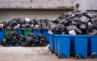 A pile of overflowing black garbage bags spilling over the top of several blue and green recycling...