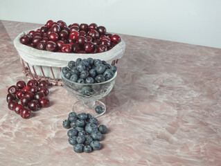 a lot of blueberries and cherries in a glass vase and in a basket are on the table for natural nutrition.