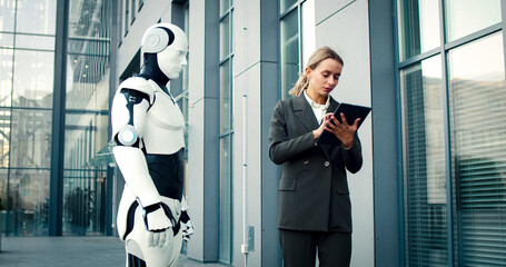 Portrait of Caucasian female wearing business suit stands near robot. Businesswoman make report on...