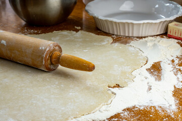 Making a pie.   Dough on a floured wooden surface.  Side view includes rolling pin, prepared sliced apples, white pie plate, butter and a wooden rolling pin.