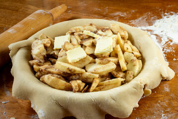 Making a pie.   Rolled dough placed over a pie shell and filled with sliced apple filling with butter.  Top side view.  Rolling pin in background.