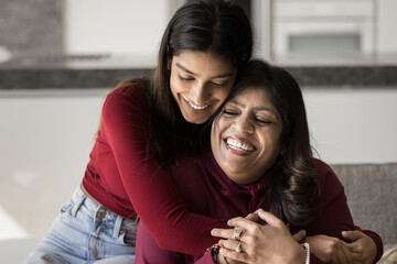 Happy beautiful young Indian daughter woman embracing senior mom from behind with love, tenderness. Cheerful mother and adult kid hugging with closed eyes on home couch, smiling, laughing