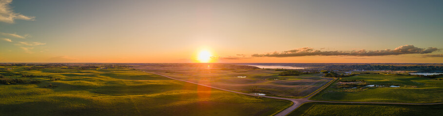 A wide aerial panoramic view of the sun setting over a vast prairie agricultural landscape that is cut by gravel roads.  The setting sun highlights the rolling farm fields.
