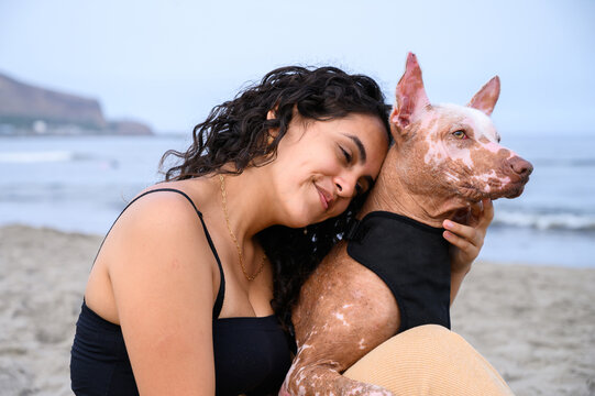 Young woman is cuddling her senior xoloitzcuintli dog on a beach, enjoying a tender moment of