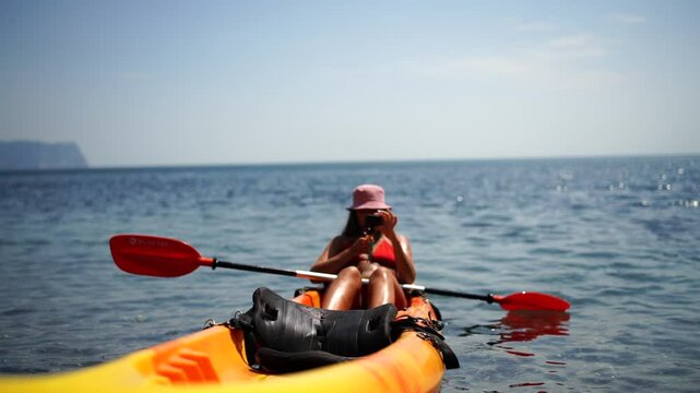A woman is sitting in a kayak on a sea. She is wearing a red and yellow life jacket and a pink hat. She is looking at her phone while paddling.