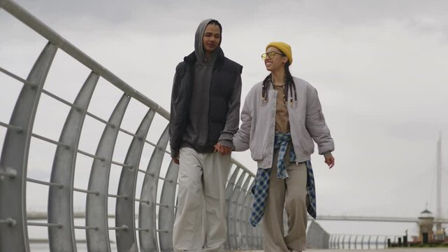 Full shot of young happy Biracial couple wearing hipster clothes talking and holding hands while having date outdoors, walking on promenade and enjoying time together