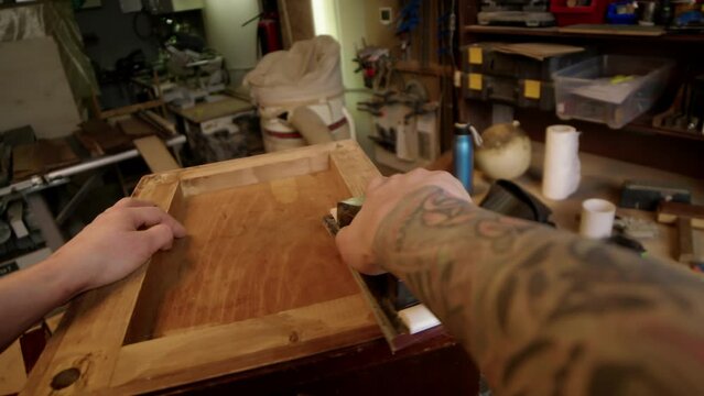 POV of tattooed male carpenter using manual sander while cleaning and polishing piece of old wooden furniture at workbench in workshop