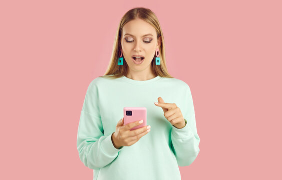 Portrait of amazed joyful blondy girl with long hair in mint sweatshirt looking at smartphone screen aand pointing finget to phone on pink background. Online shopping, browsing social media concept.